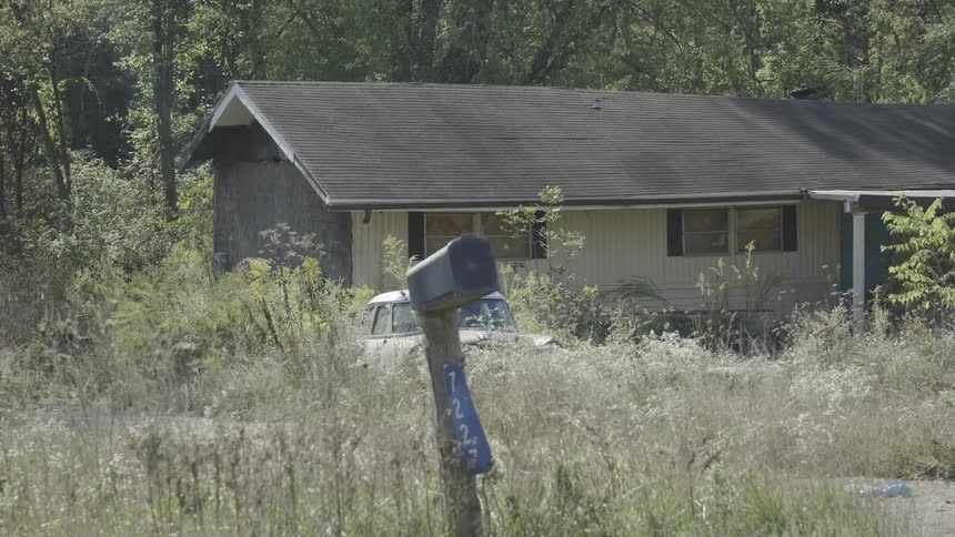 A slow zoom out of an old, abandoned rural house with an antique car parked in the overgrown front yard