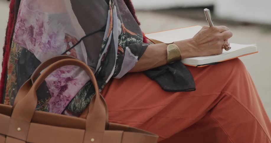 Close up on the hands of a stylish woman as she sits outdoors writing her thoughts in a journal