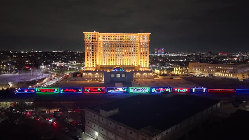 A brightly illuminated holiday train passes in front of a grand historic building in a bustling city at night