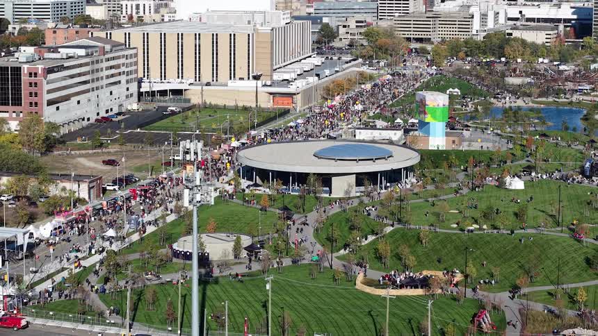 Slow-pulling aerial view captures a massive crowd at an outdoor festival in a downtown Detroit park on a sunny day, revealing the city skyline