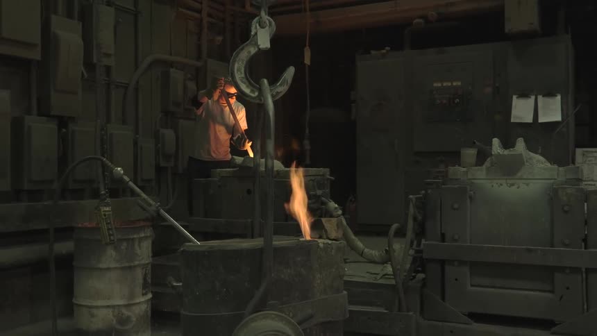 An industrial worker in a foundry tends to molten metal as a flame burns in the foreground