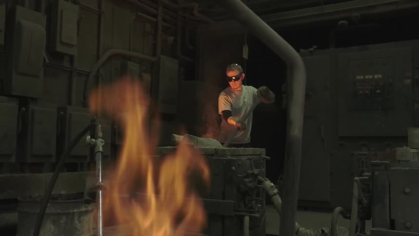 A factory worker in protective gear stirs molten metal in a crucible with a fire burning in the foreground