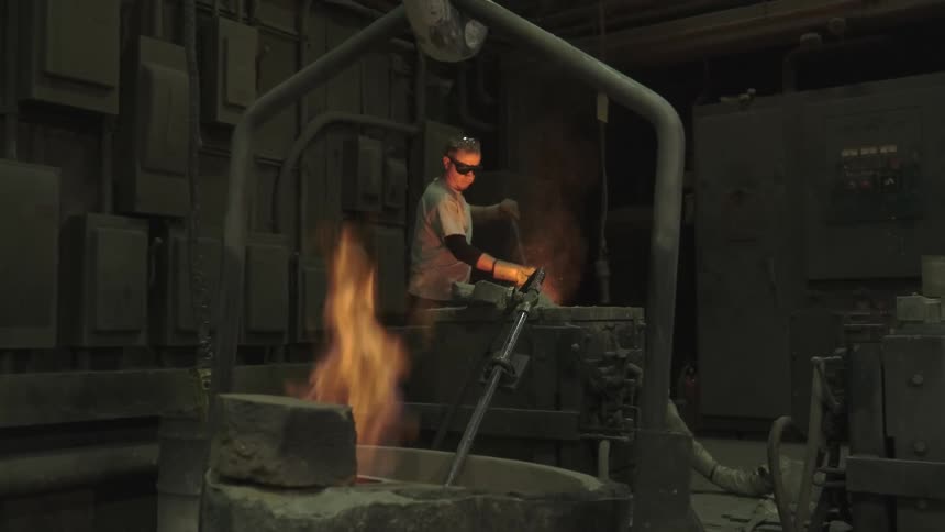 A factory worker in protective gear tends to a vat of molten metal as flames burn in a crucible in the foreground