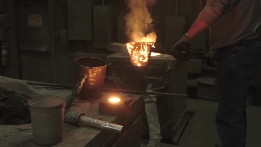 A foundry worker carefully moves a ladle of glowing hot molten metal in an industrial factory setting