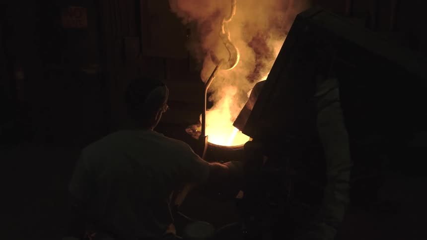 Industrial workers in a dark foundry pour glowing molten metal into a crucible in slow motion