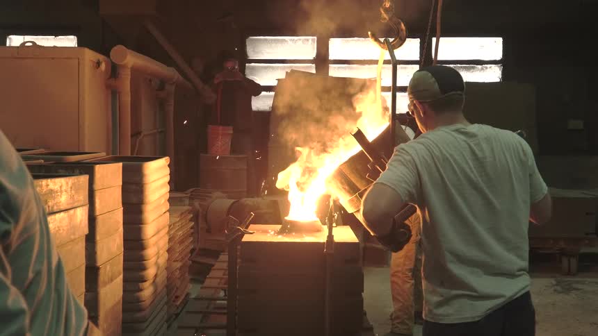 A team of factory workers carefully pour glowing hot molten metal into a casting mold in slow motion