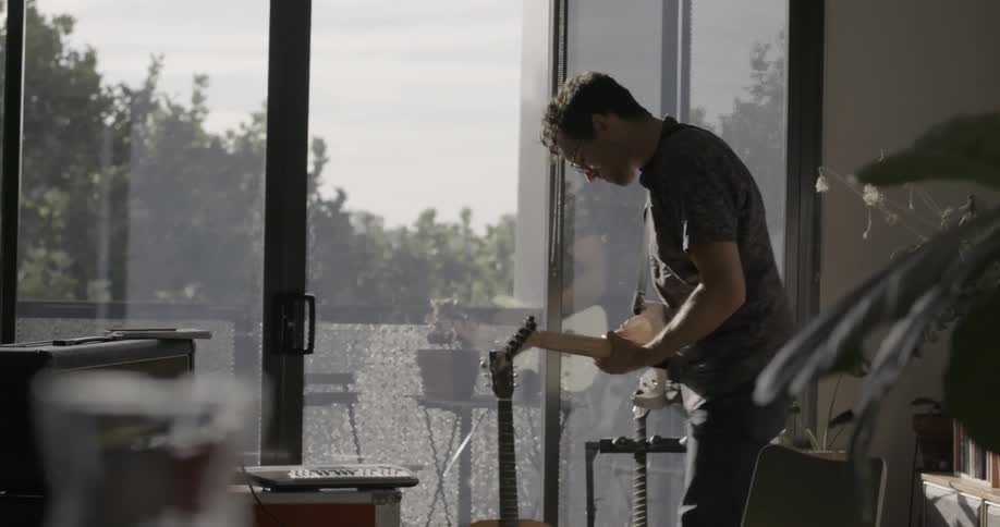 A man plays guitar in front of a large window during the day