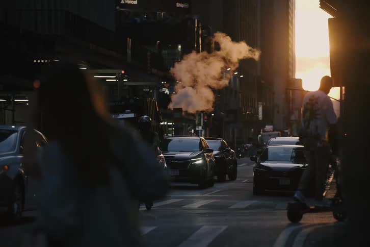 Cinematic slow-motion shot of a bustling city street at sunset, with steam rising into the golden light as traffic and pedestrians pass by