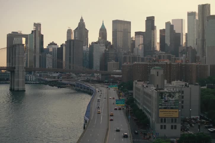 A view of the Lower Manhattan skyline and the Brooklyn Bridge through a fence, with traffic moving along the FDR Drive at sunset