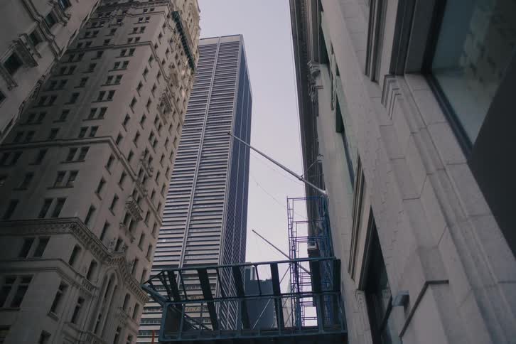 A low-angle dolly shot pulls back from towering skyscrapers to reveal street-level scaffolding in a city financial district