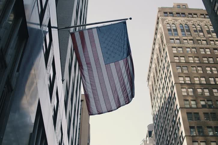A slow-motion, low-angle shot of an American flag hanging between skyscrapers, tilting up to reveal two military jets flying overhead
