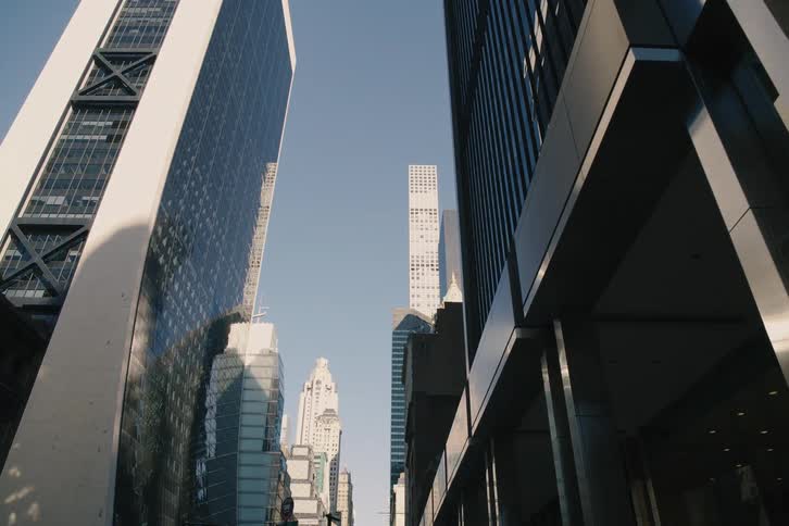 A slow dolly shot moves down a sunny New York City street canyon, revealing towering skyscrapers, classic architecture, and a prominent American flag