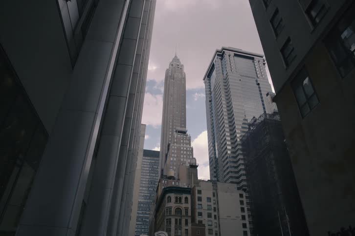 A slow dolly out from a low angle reveals towering skyscrapers reaching for the cloudy sky in a dense city canyon