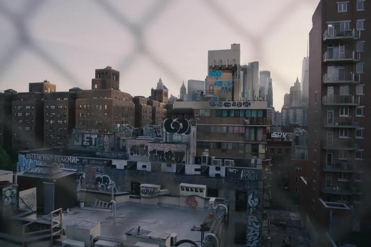 A slow, contemplative pan across graffiti-covered rooftops in a dense New York City neighborhood, viewed through a chain-link fence at dusk