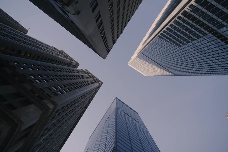 A slow, dramatic low-angle view reveals towering skyscrapers reaching towards a clear, vast sky in a bustling downtown district