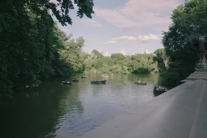 A scenic view from a stone bridge of people leisurely rowing boats on the lake in Central Park on a sunny day