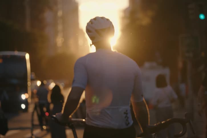 A cyclist in full gear stands on a busy city street, backlit by the warm, golden glow of the setting sun
