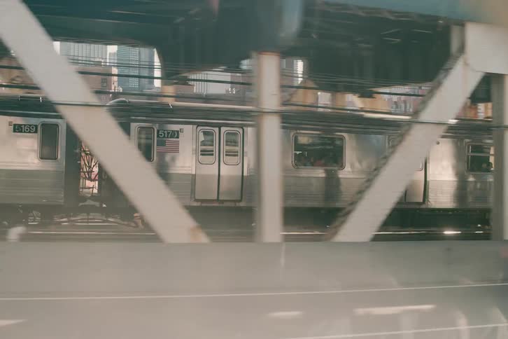 A cinematic tracking shot from a car captures a subway train crossing an industrial bridge, with sunlight flaring through the girders