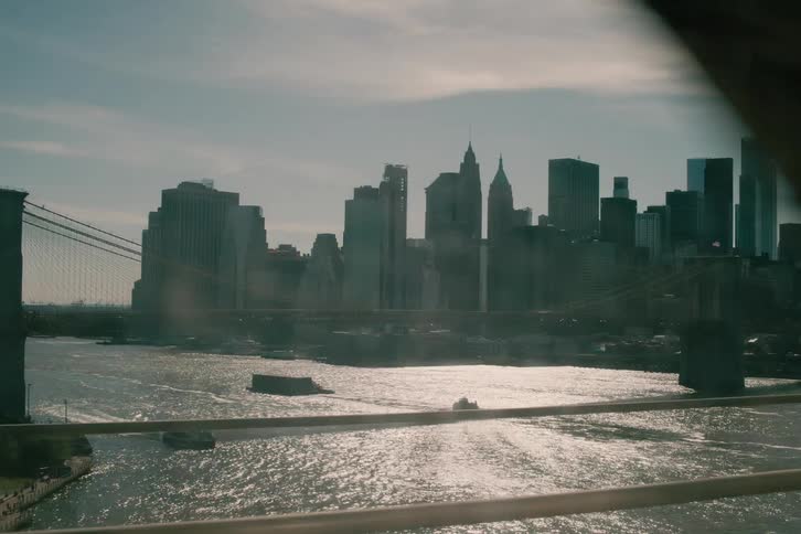 A cinematic tracking shot of the sunlit Lower Manhattan skyline and Brooklyn Bridge as seen from a moving train