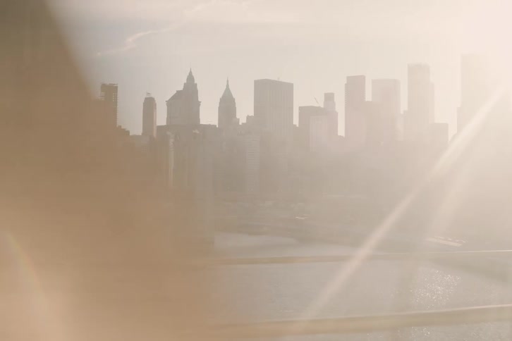 A dynamic time-lapse view from a moving train reveals the Lower Manhattan skyline and Brooklyn Bridge bathed in a dreamy, hazy sun flare