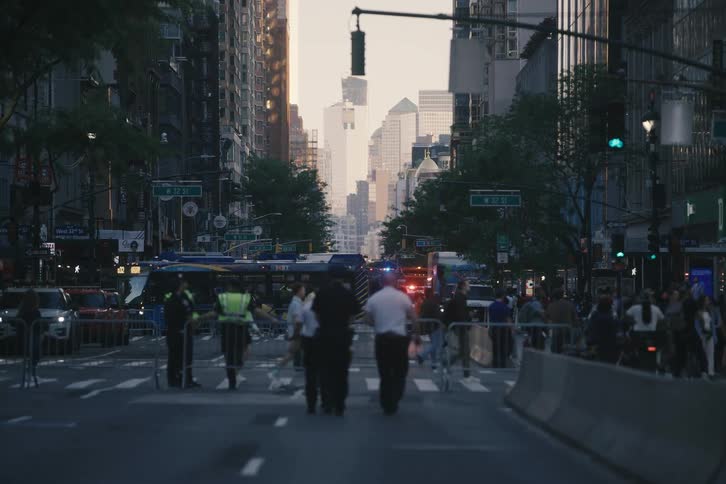 Police officers stand guard on a blocked-off New York City street at dusk with barricades and crowds in the background
