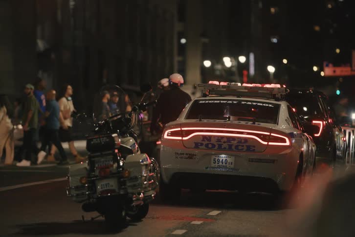 An NYPD highway patrol car and a motorcycle officer with flashing lights are parked on a busy New York City street at night