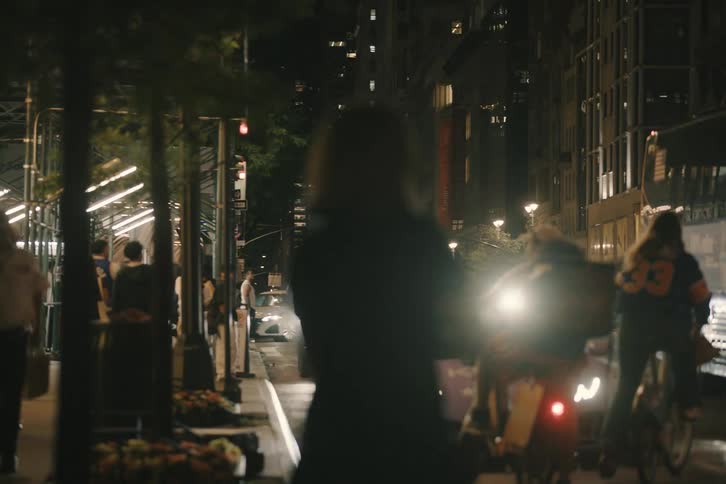 A woman stands alone in the middle of a busy city street at night, observing the traffic and city lights
