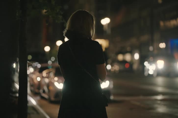 A solitary woman stands on a rainy city sidewalk at night, watching the traffic and bright lights pass by