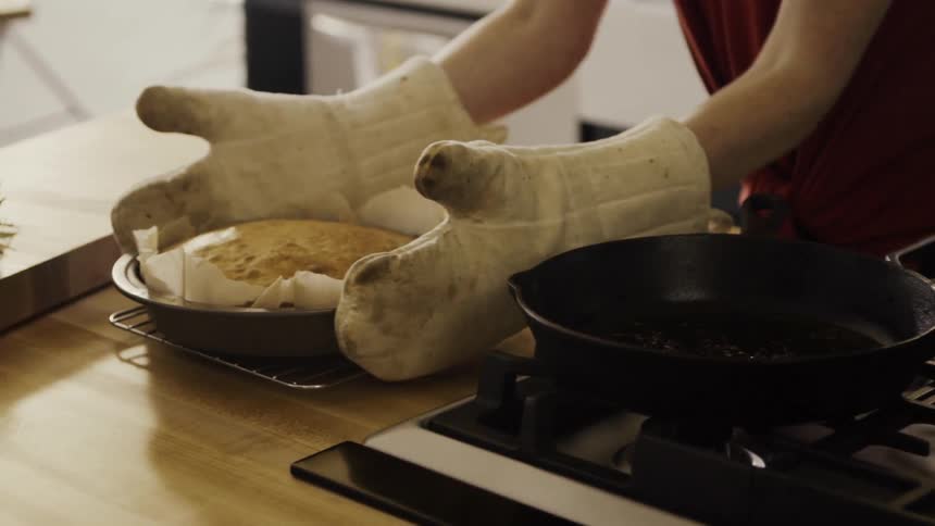 A woman wearing mitts places a baked cake on a cooling rack