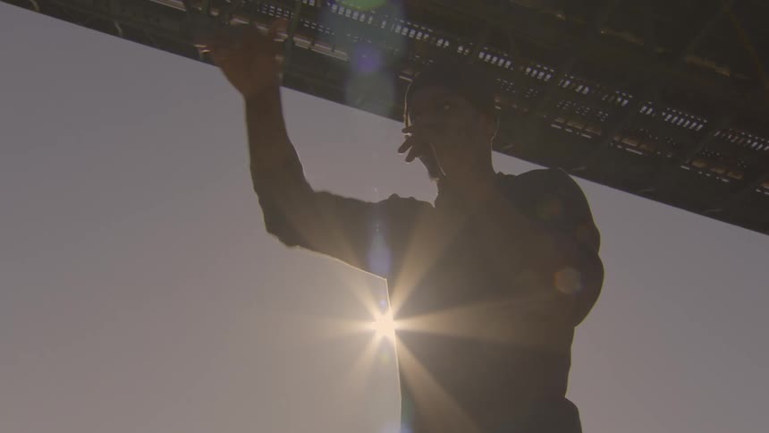 A silhouetted young man with braids performs an expressive slow-motion dance while playing a harmonica under a city bridge at sunset