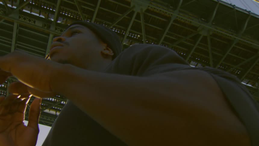 A contemplative young man looks into the distance while standing underneath a large industrial bridge during golden hour