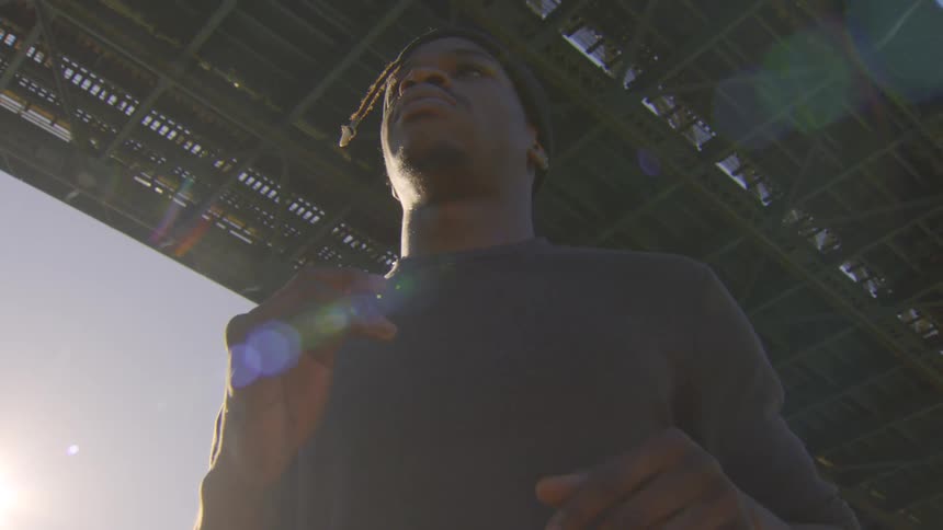 A low-angle slow-motion shot captures a determined man jogging in place under a large bridge with bright sunlight flaring into the camera