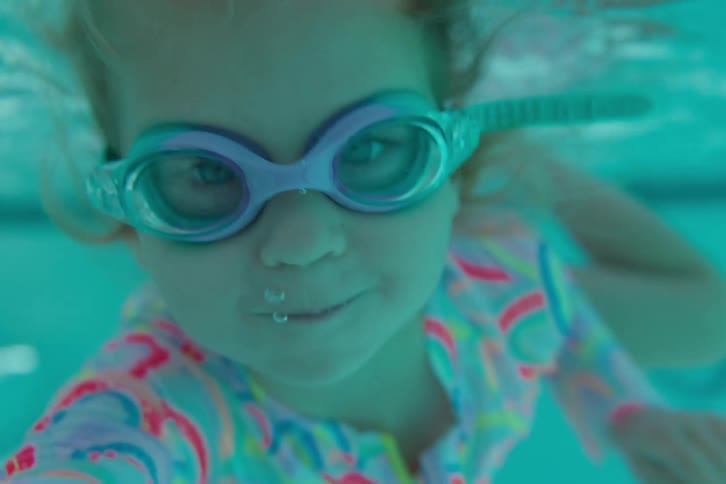 A smiling young girl in goggles swims towards the camera underwater before surfacing in a pool