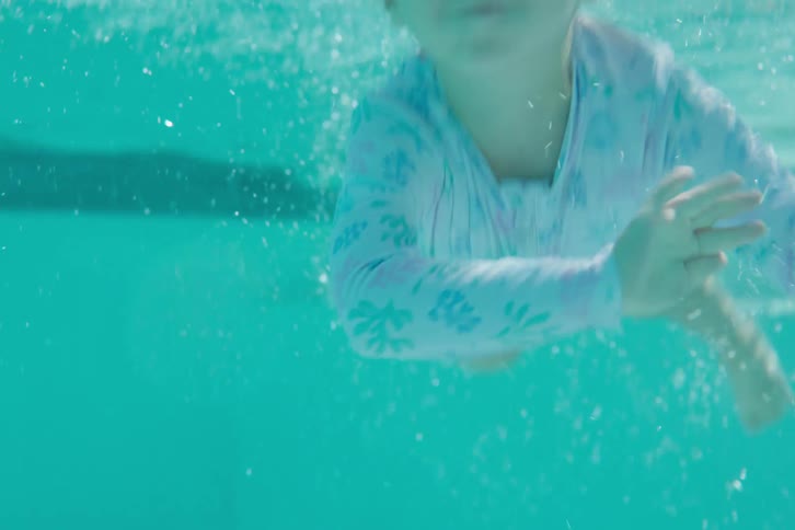 A young girl with goggles jumps into a swimming pool and swims towards the camera underwater in slow motion