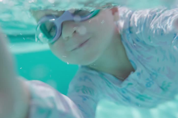 An immersive underwater point-of-view of a young child with goggles playfully swimming in a turquoise pool