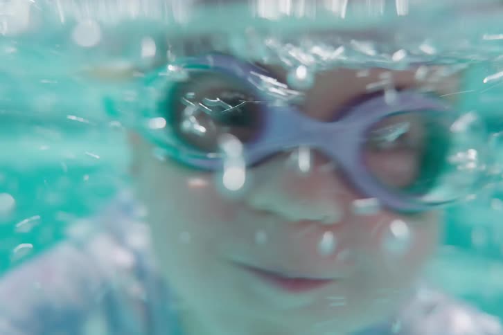 A playful little girl in goggles dunks the camera underwater for a fun, bubbly, first-person view in the swimming pool