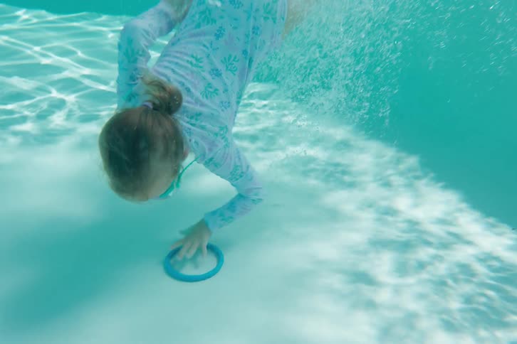 An adventurous little girl jumps backwards into the pool and swims underwater to retrieve a diving ring in slow motion