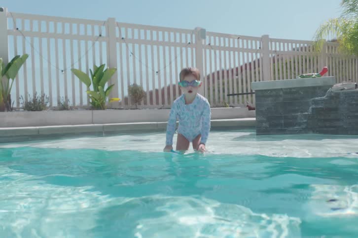 A young girl in goggles plays with rings in a pool before swimming towards the camera, with a slow-motion underwater view