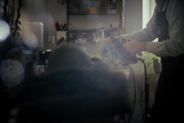 A hatter steams the brim of a felt hat with an iron in his dimly lit workshop, viewed from across the room