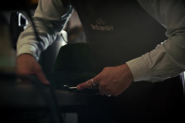 An artisan hat maker meticulously shapes the brim of a green felt hat in a dimly lit workshop