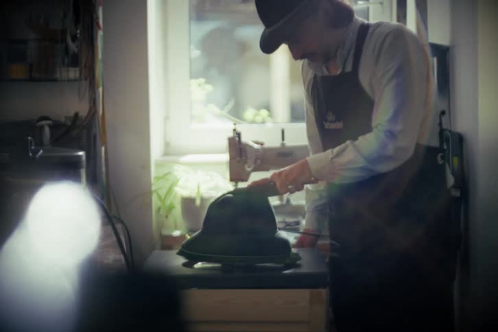 A skilled hatter carefully brushes and shapes a felt hat on a spinning block in his sunlit workshop