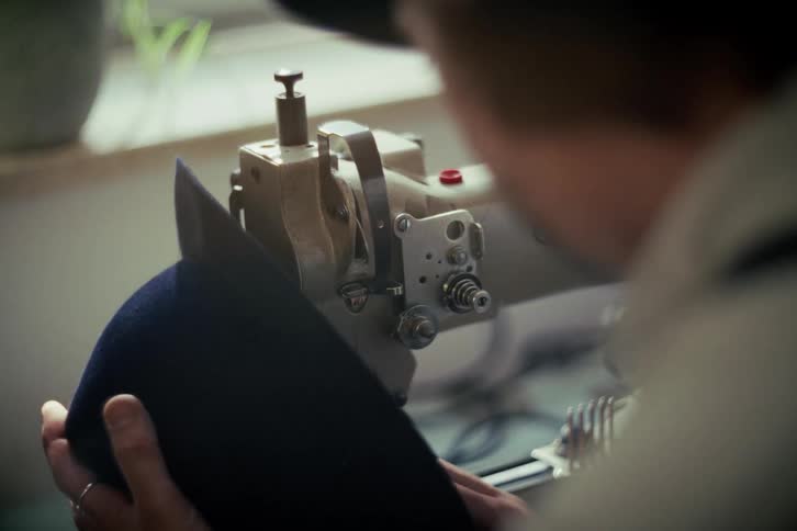 Over the shoulder shot of a skilled hat maker using an industrial sewing machine to carefully craft a cap in a workshop