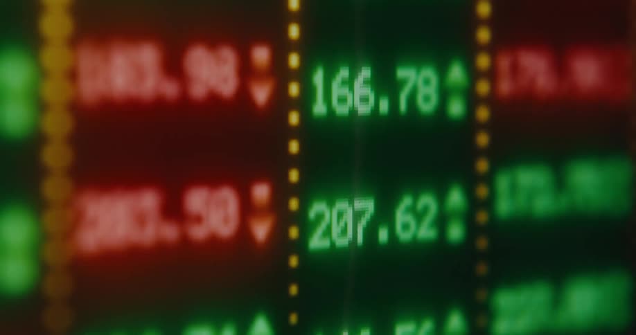 A soft-focus close-up of a digital stock market board shows fluctuating red and green numbers, symbolizing market volatility and trading