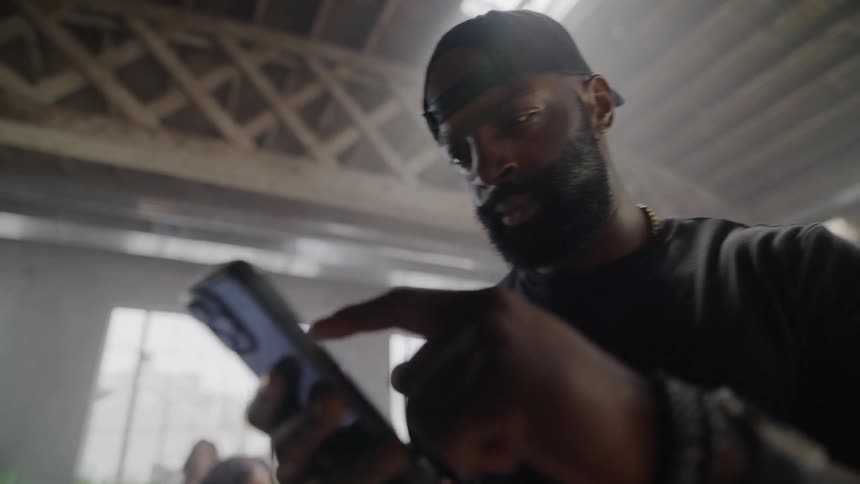 Low angle shot of a focused man in a backward cap scrolling on his smartphone inside a hazy industrial loft