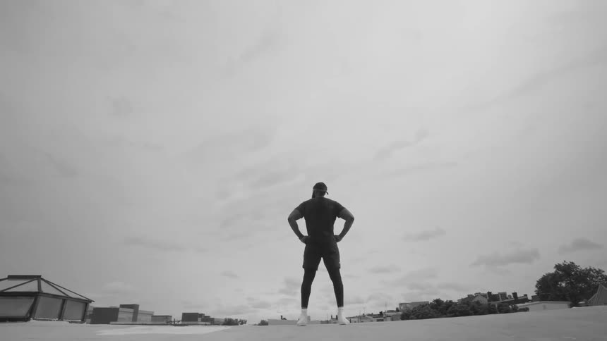 Dramatic black and white slow-motion shot of a muscular man standing in a power pose on a city rooftop, looking out at the vast sky