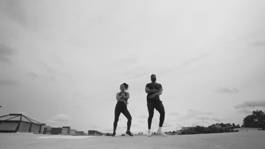 A man and a woman perform a synchronized, energetic dance routine in slow motion on a city rooftop in black and white
