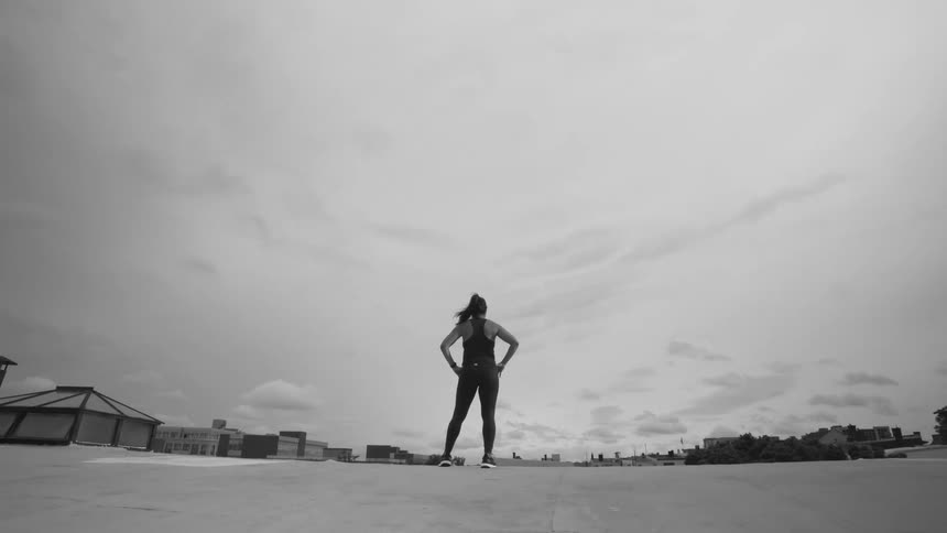 A powerful athletic woman stands with her hands on her hips on a city rooftop against a vast, cloudy sky in this dramatic black and white shot