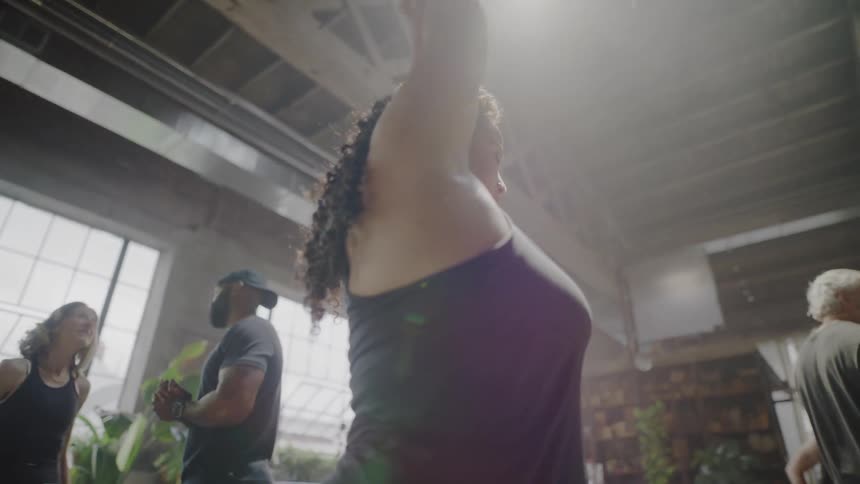 A woman with curly hair stretches her arms in slow motion during a yoga class in a bright, sunlit loft studio