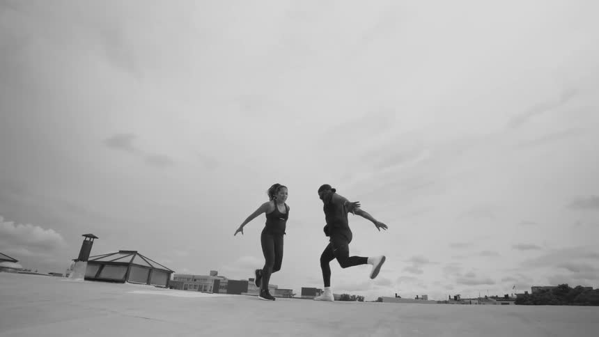 A man and woman perform an energetic dance routine in slow motion on a city rooftop in black and white
