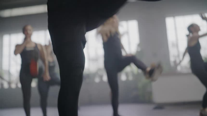 A charismatic male fitness instructor leads an intense cardio dance class in slow motion inside a hazy, sunlit gym